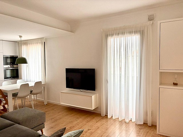 Zona de estar luminosa con mueble de televisión, cortinas blancas y vista parcial del comedor.