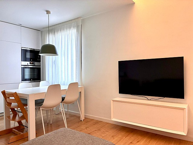Vista frontal de la cocina moderna equipada con muebles blancos, armarios superiores oscuros y mesa de comedor.